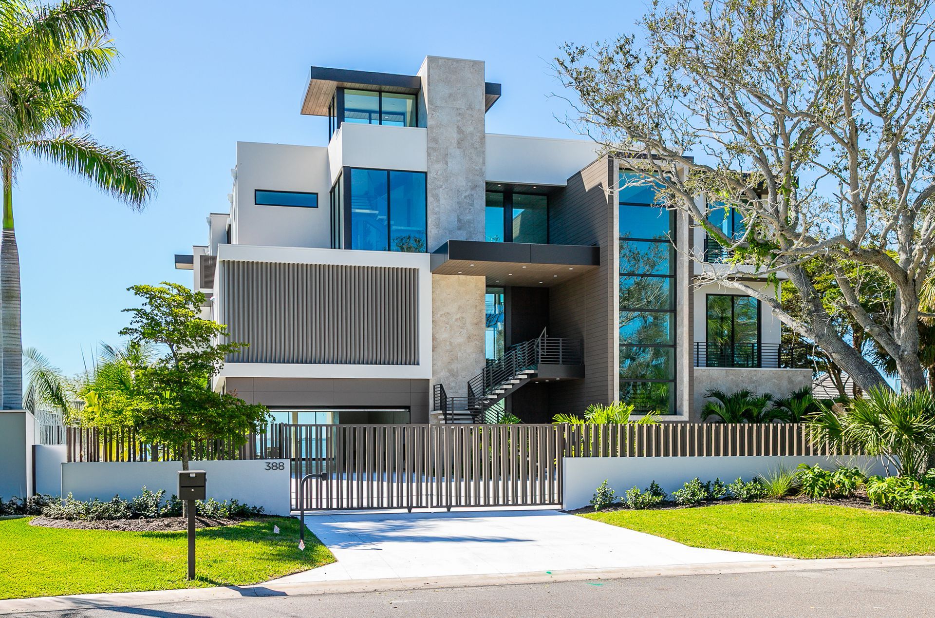 A modern three-story house with a stone facade, floor-to-ceiling windows, and a vertical wood-slat gate on a sunny day.