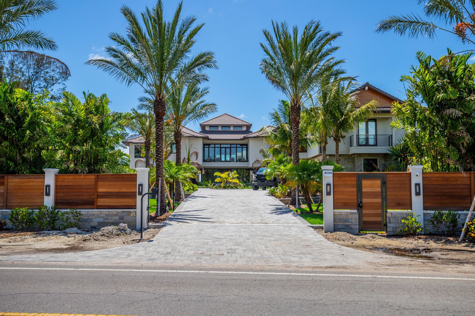 A paved driveway leads to a modern, multi-story house behind a wooden fence, framed by palm trees under a blue sky.