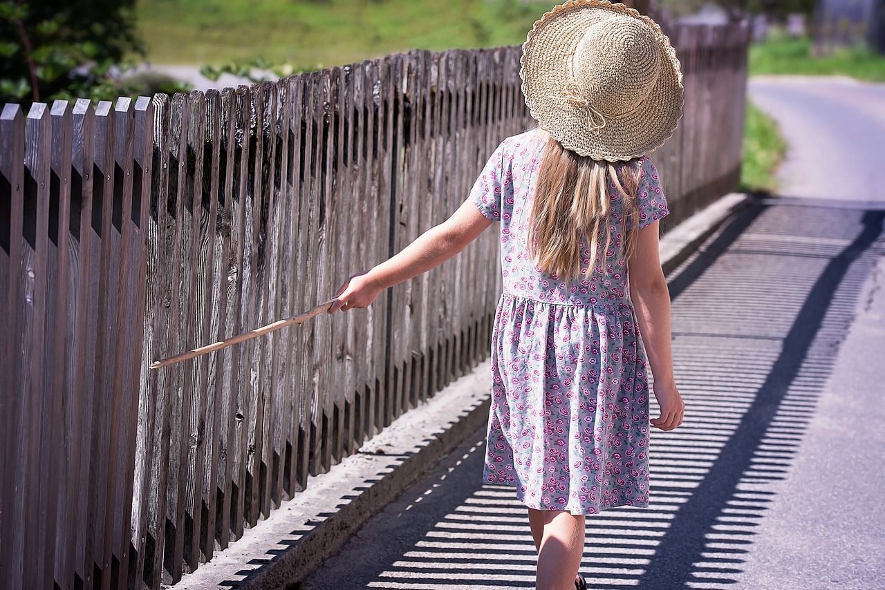 A girl walking down the street running a stick over a wooden fence