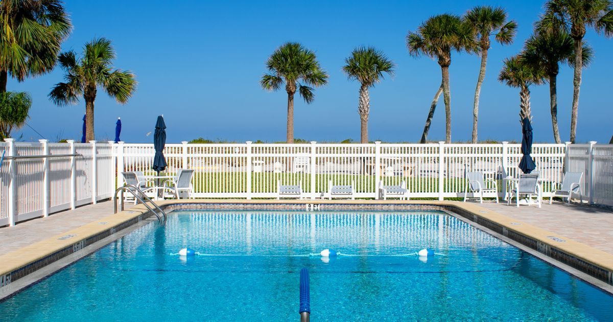 An outdoor swimming pool with blue water, surrounded by a white fence and palm trees against a clear blue sky.