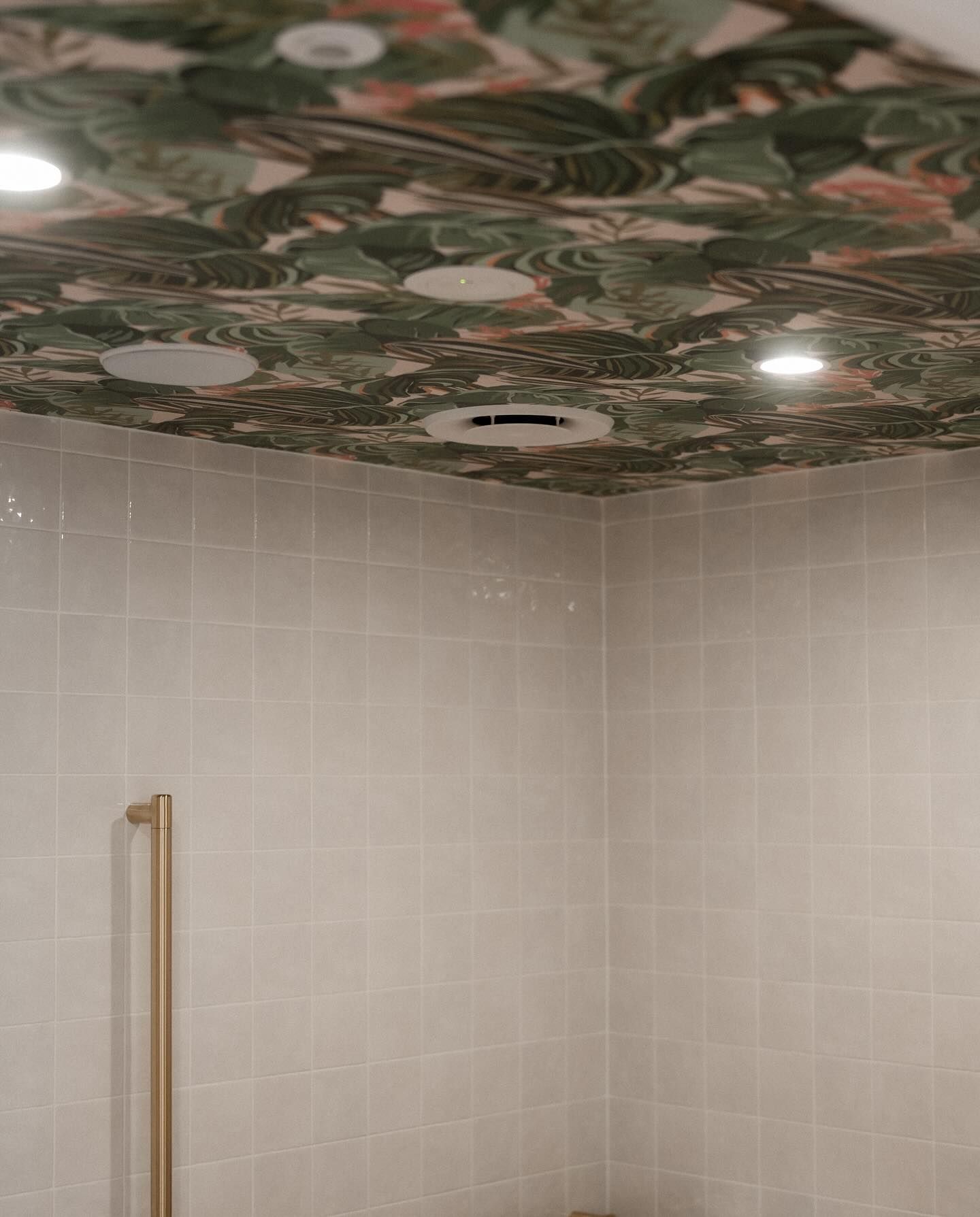 Shower interior with floral-print ceiling. Beige tiled walls, gold shower handle, recessed lights, and air vent visible.
