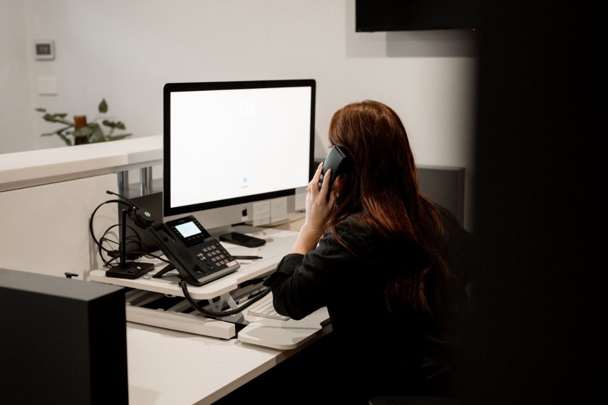 A Woman is Sitting at a Desk Talking on a Phone in Front of a Computer — John McEwan Electrical In Dapto, NSW