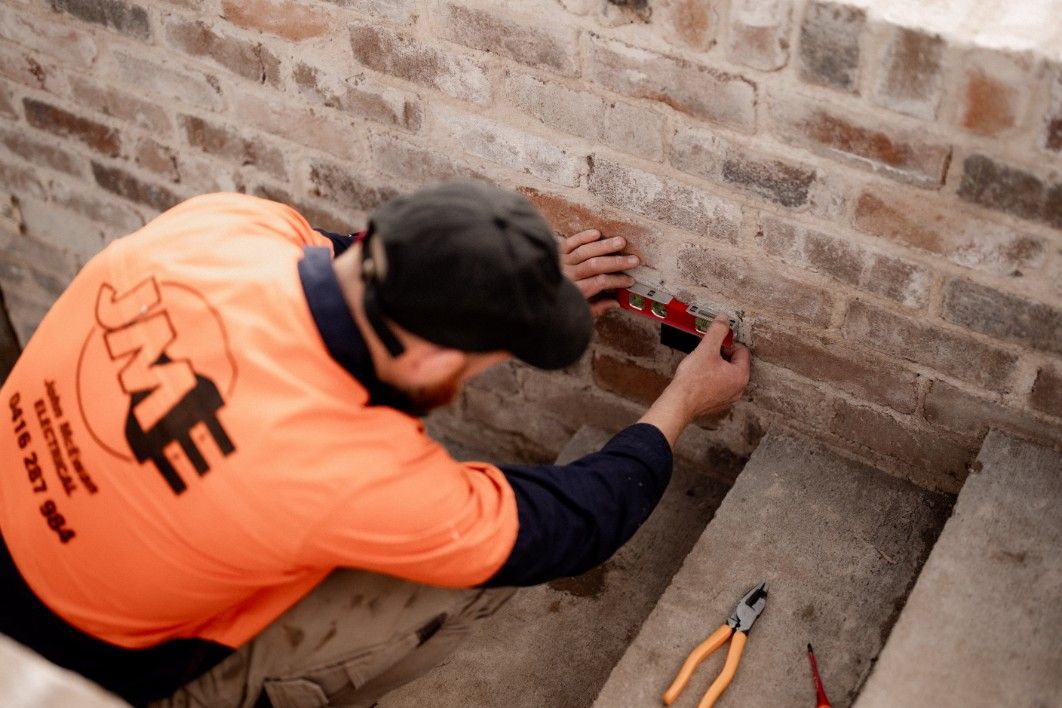 A Man is Working on a Brick Wall Wearing an Orange Shirt — John McEwan Electrical in Calderwood, NSW