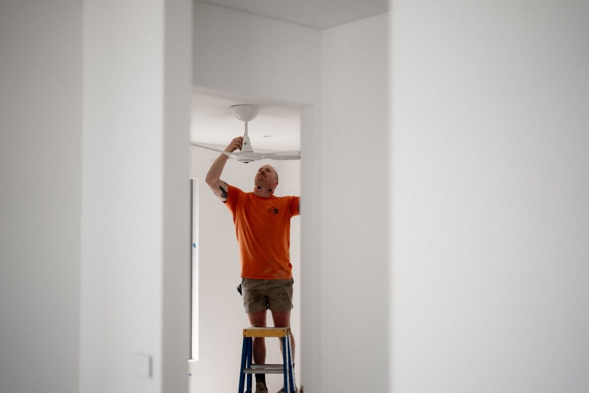 A Man is Standing on a Ladder Fixing a Ceiling Fan — John McEwan Electrical in Corrimal, NSW