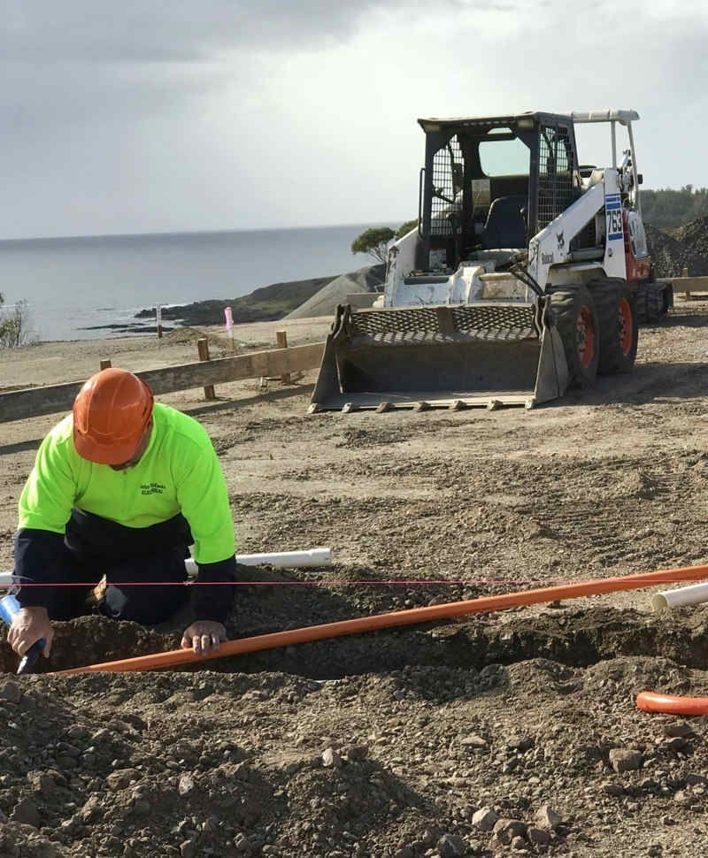 A Man Is Kneeling In The Dirt Next To A Bulldozer — John McEwan Electrical In Dapto, NSW