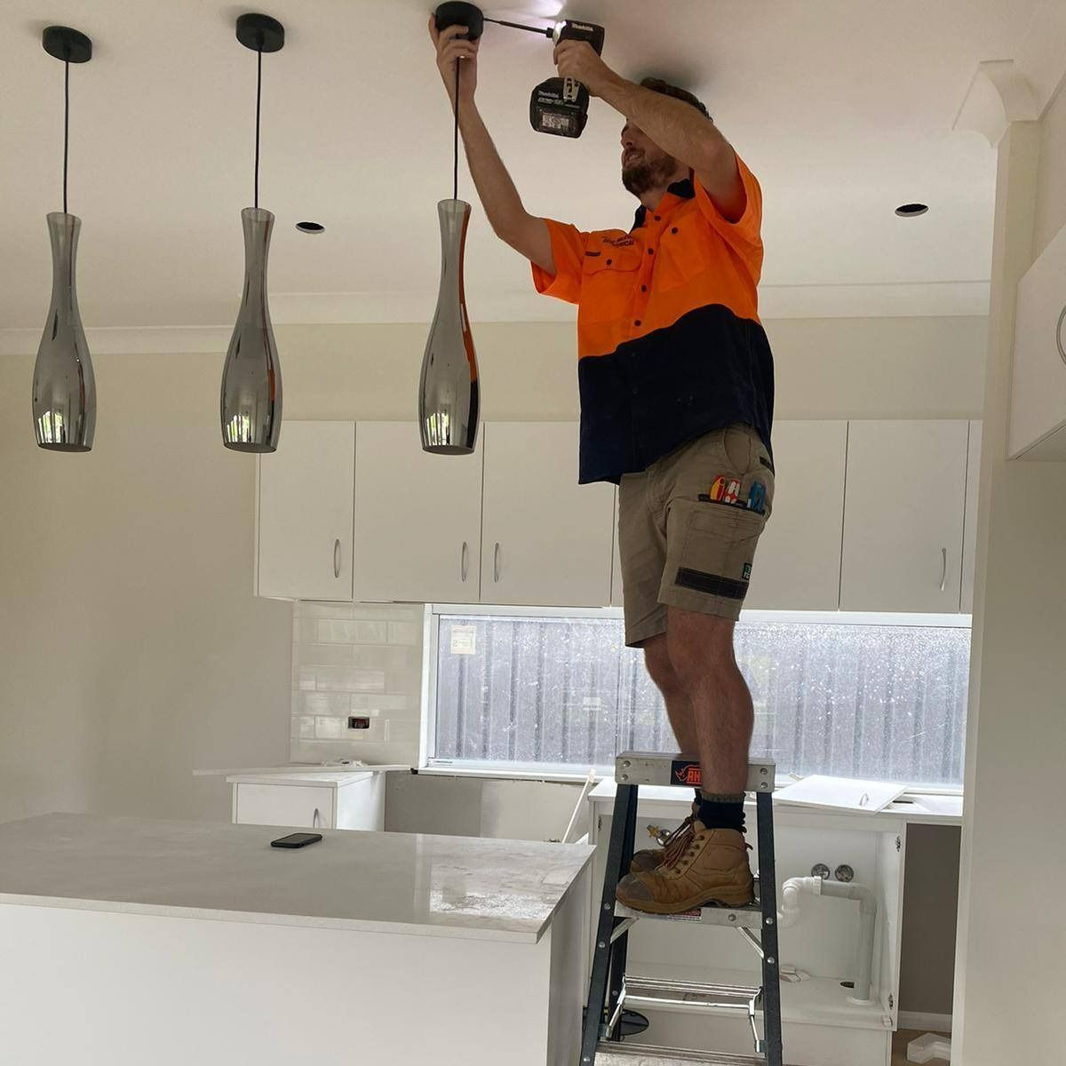 A Man Is Standing On A Ladder Working On A Light Fixture — John McEwan Electrical In Unanderra, NSW