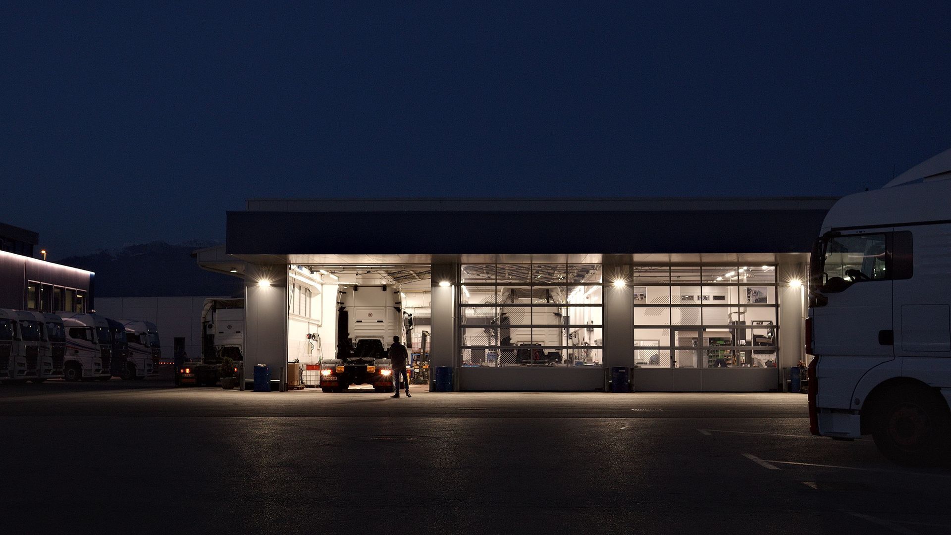 A mechanic works late in a well-lit truck repair shop, surrounded by parked trucks.
