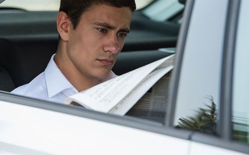 A man is sitting in the back seat of a car reading a newspaper.