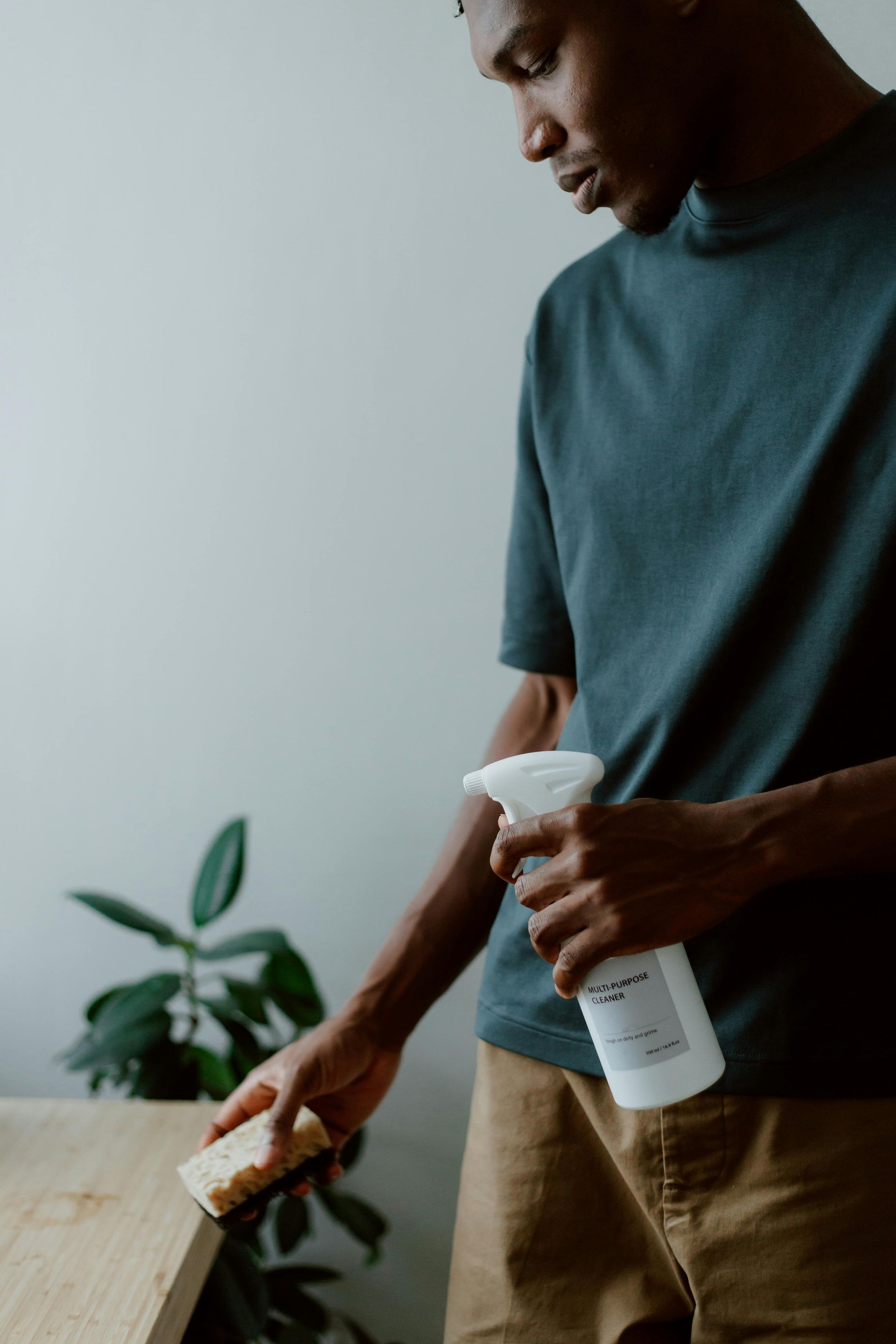 A man is cleaning a wooden table with a spray bottle and a sponge.
