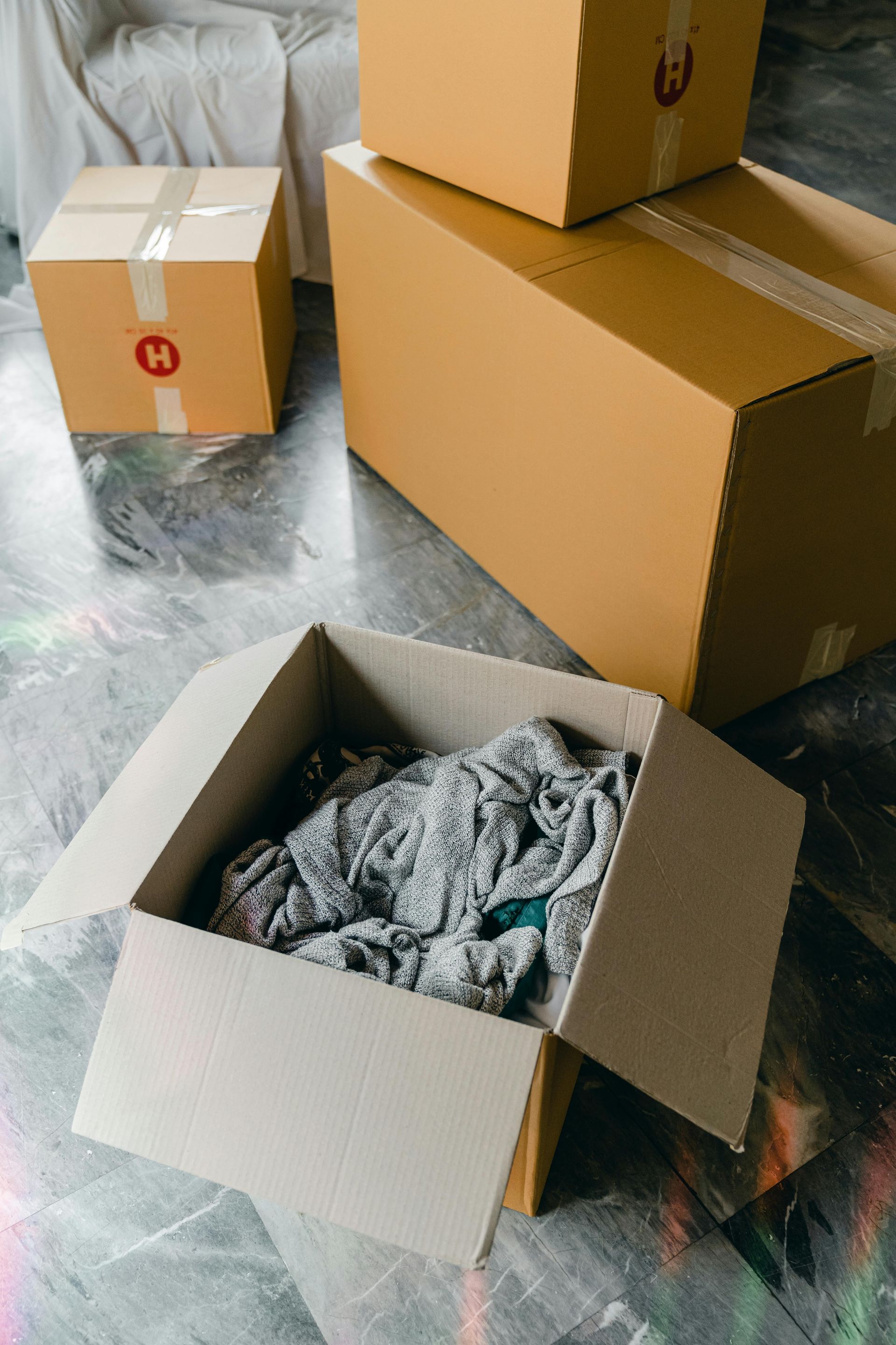 A cardboard box filled with clothes is sitting on the floor next to a stack of cardboard boxes.