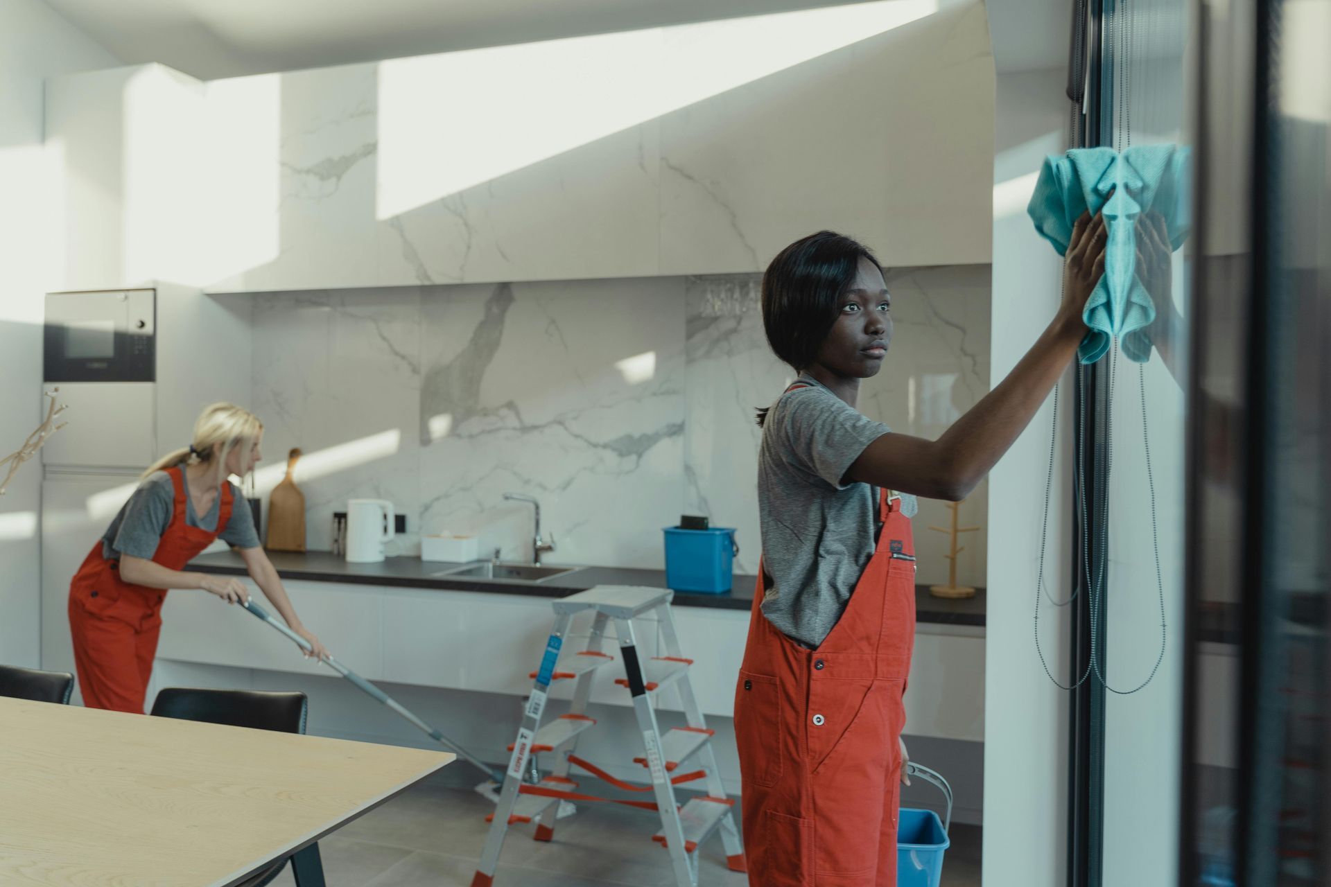 Two women are cleaning a window in a kitchen.