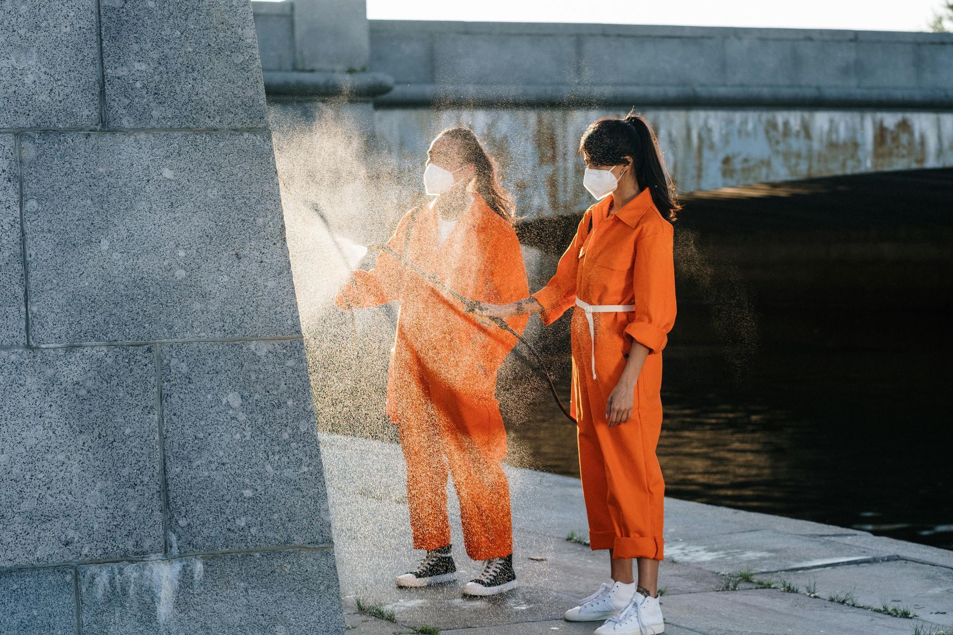 Two women in orange jumpsuits and masks are spraying powder on a wall.