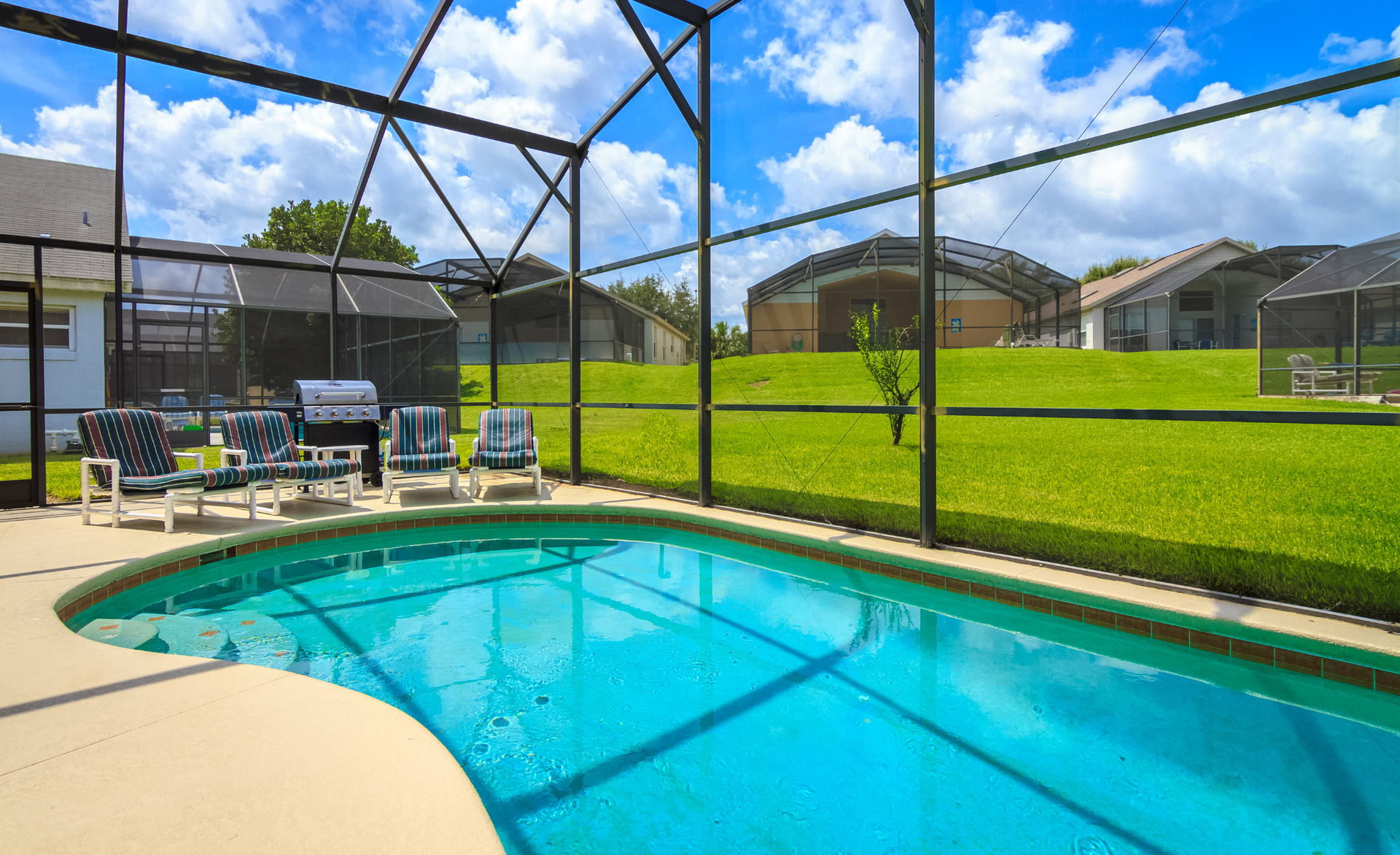 A large swimming pool surrounded by a screened in area with chairs and a grill.