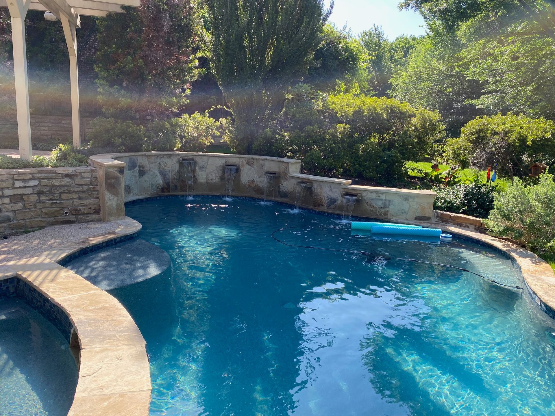 A large swimming pool is surrounded by trees and a pergola.