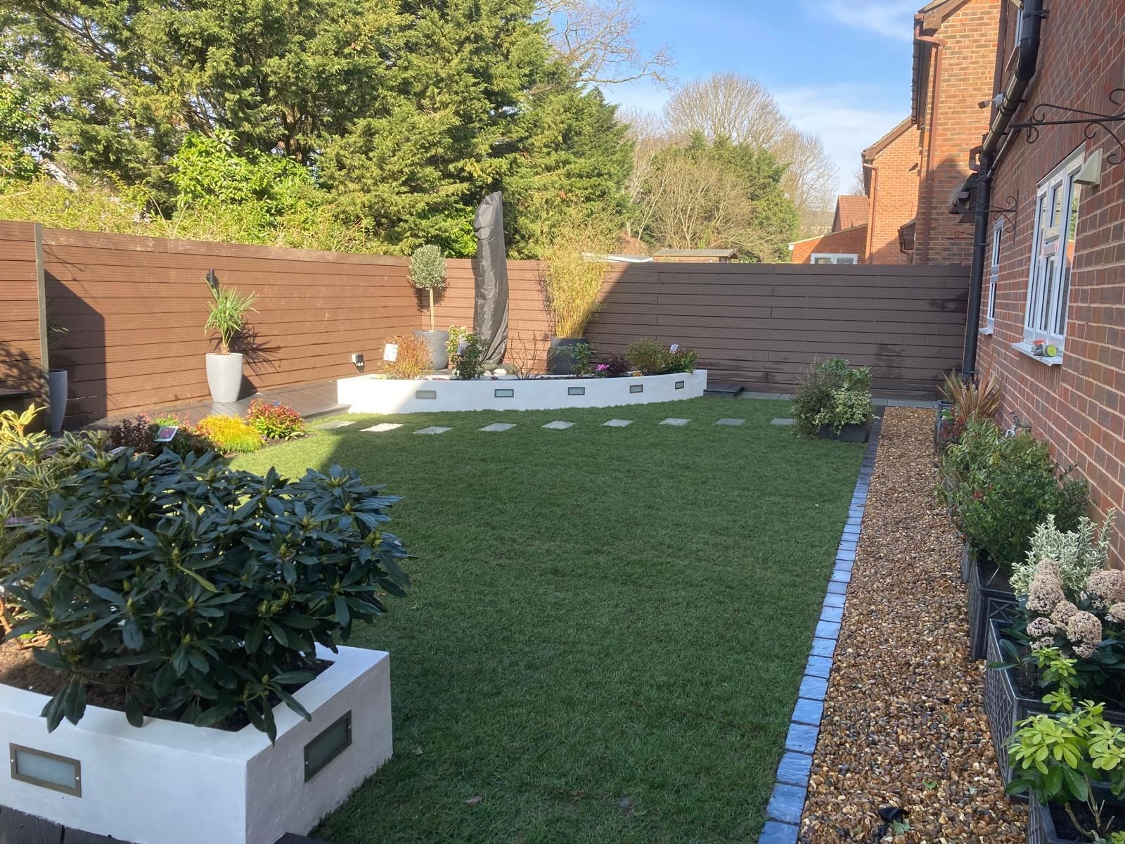 Well-kept backyard with green lawn, stone pathway, and brown wooden fence. White planters hold plants and flowers.