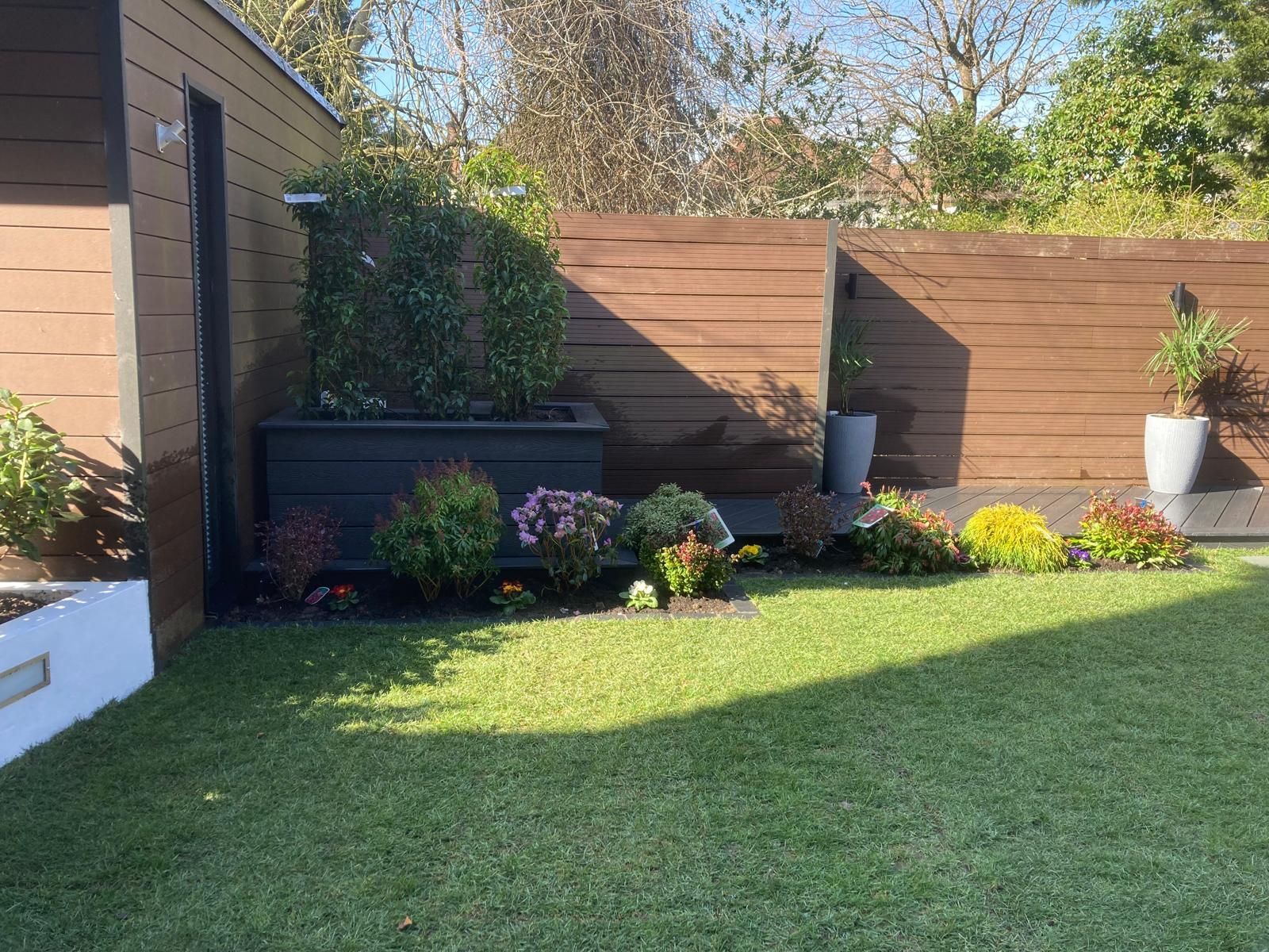 A backyard garden with a wooden fence, flower beds, and a lush green lawn. The sun casts shadows.