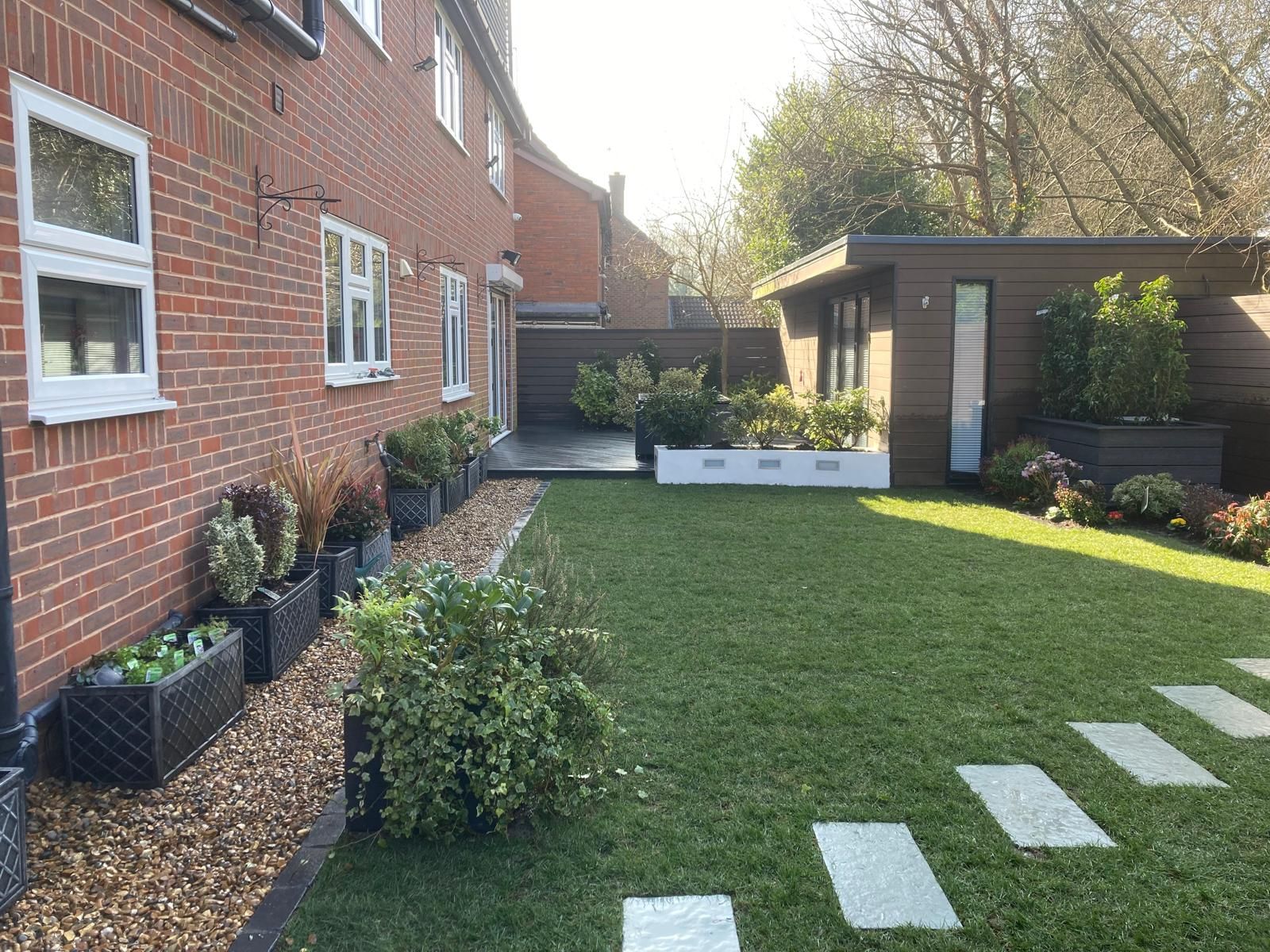 A well-manicured backyard with a brick house on the left and a modern shed at the back. The yard has a green lawn, flower beds, and a stone pathway.