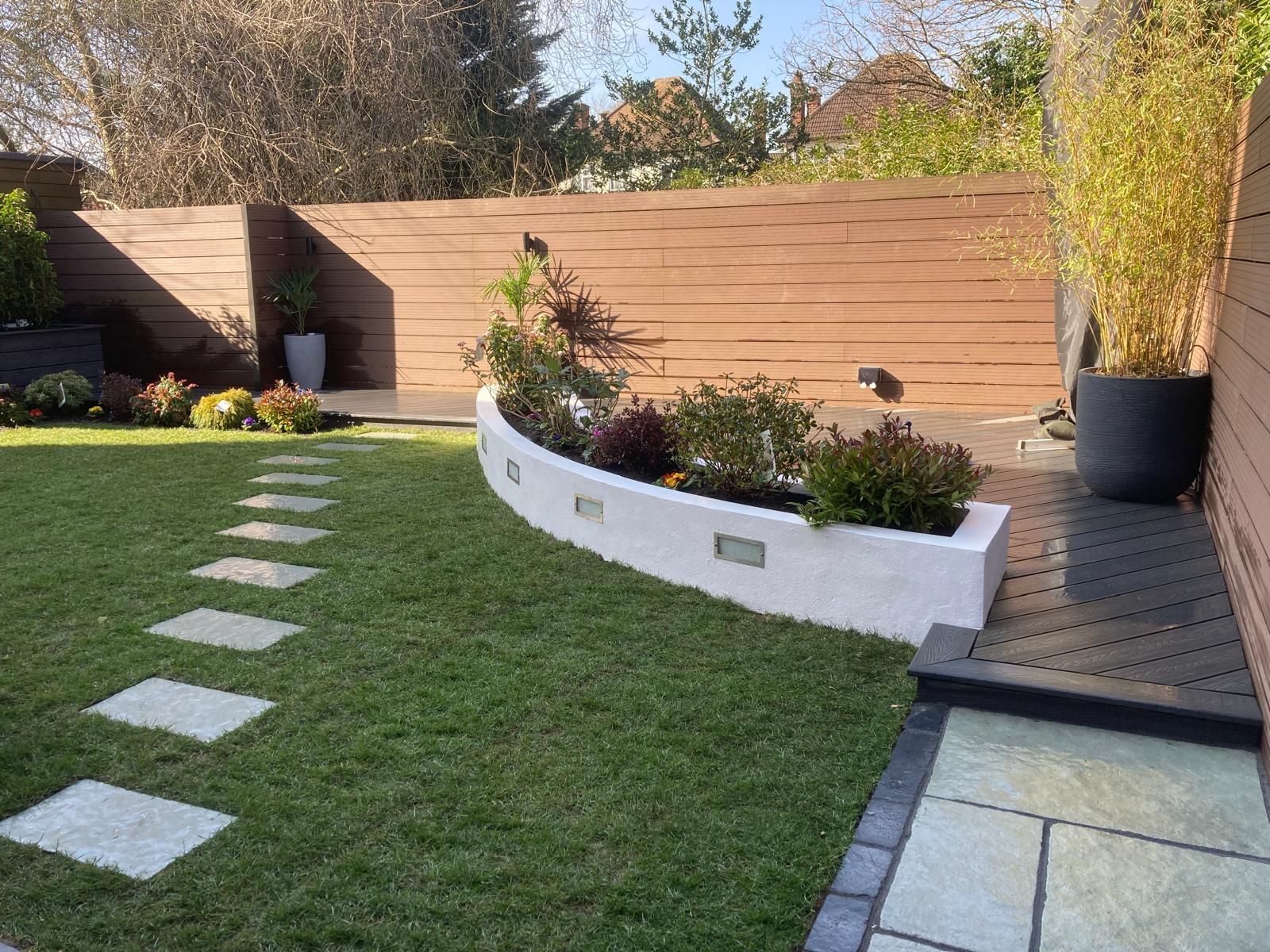 A modern backyard with a stone pathway through green grass, leading to a curved planter with low-lying shrubs. A wooden fence and black potted bamboo are in the background.
