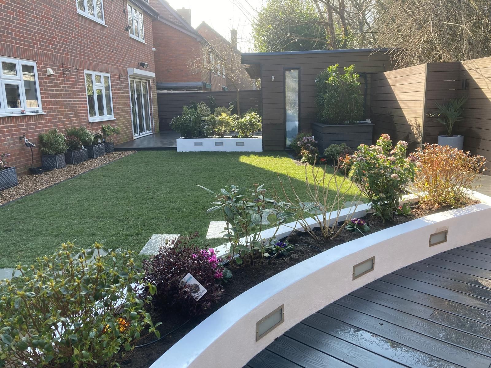 A well-manicured backyard with a green lawn, flower beds, and a wooden shed. A brick house is on the left.