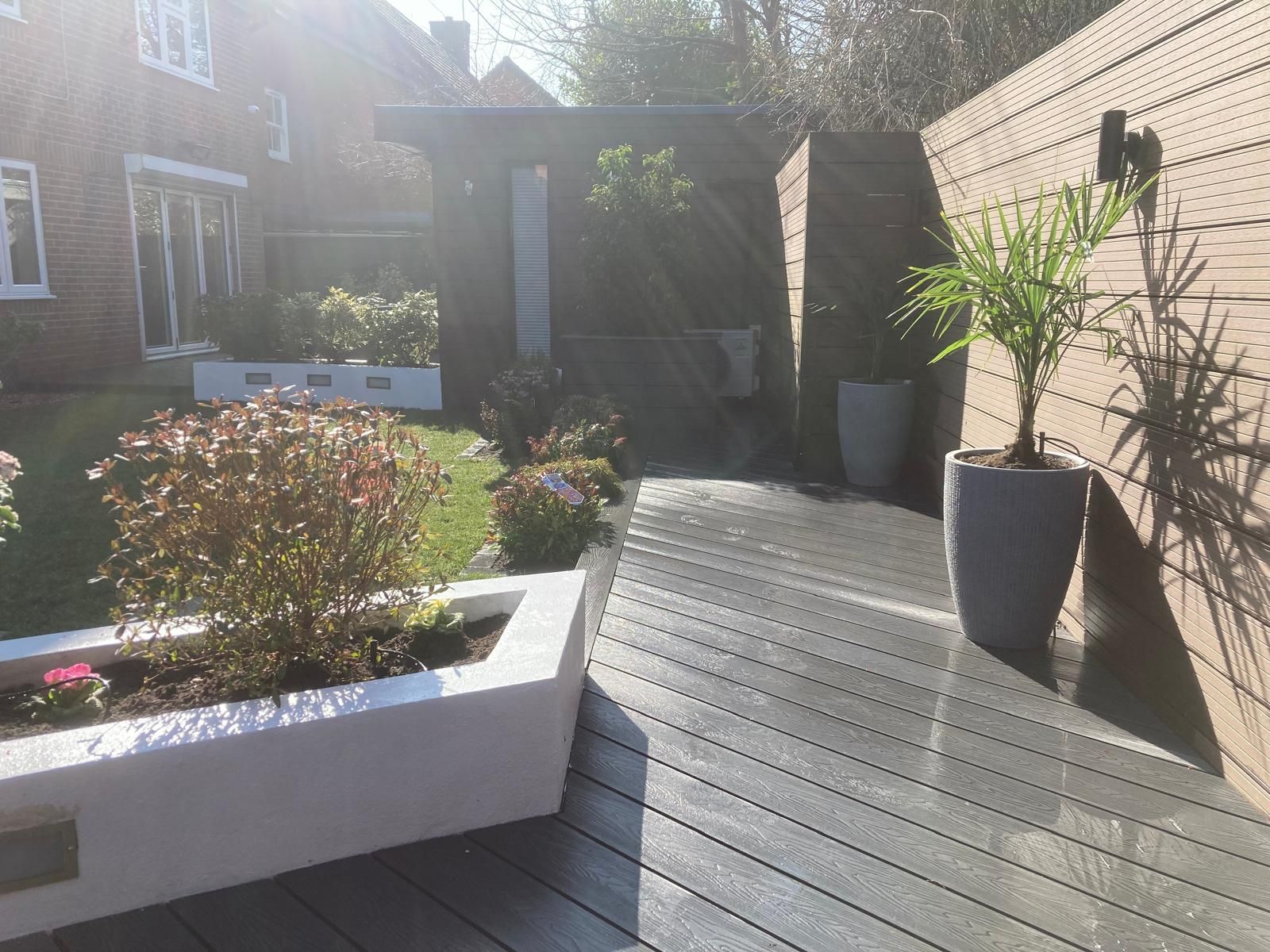 A modern garden with a composite deck, planters, and a wooden pergola. A potted palm tree stands beside a wooden fence.