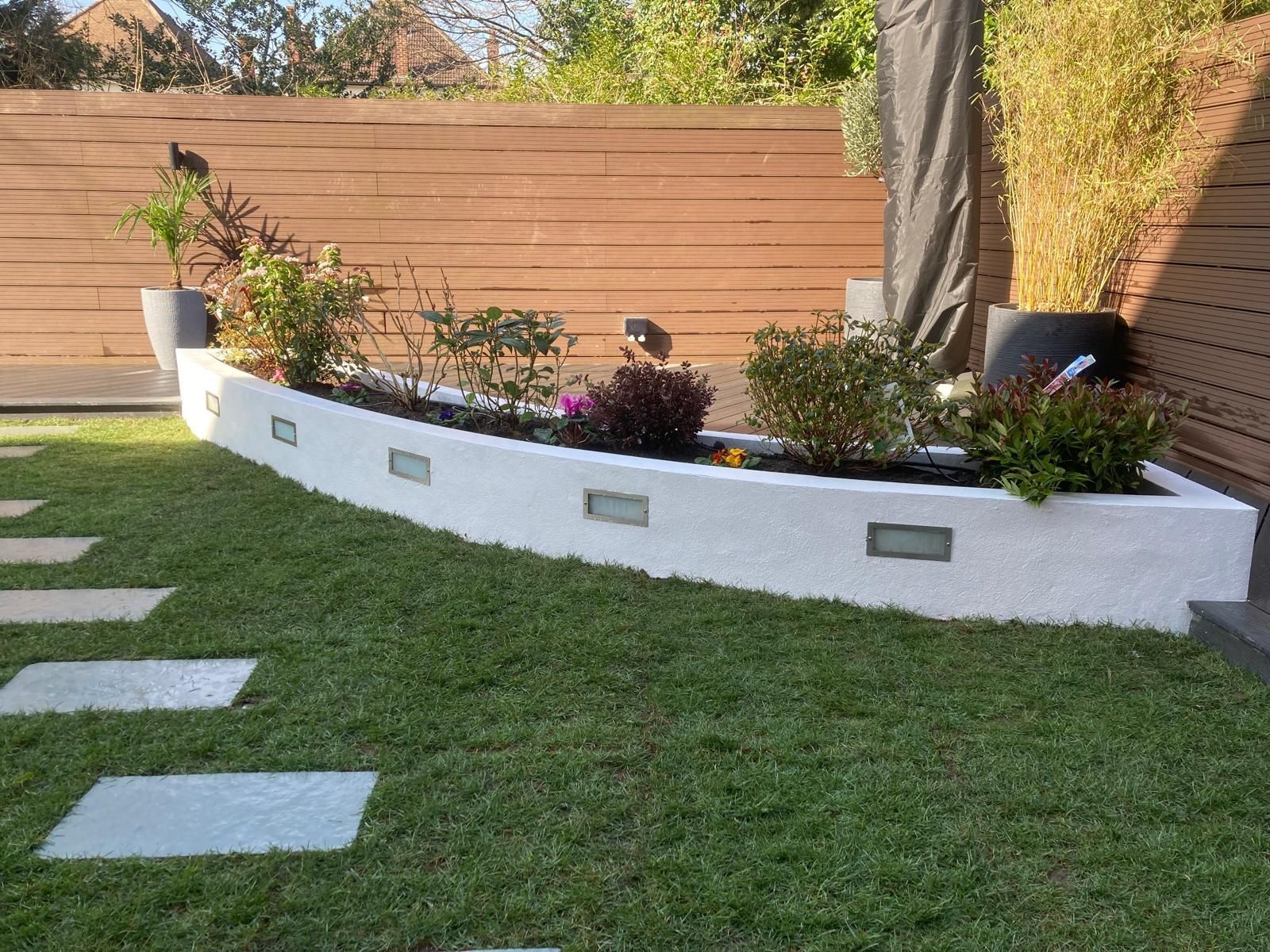 Curved white planter box with greenery and flowers, adjacent to a lawn with stepping stones and a wooden fence.