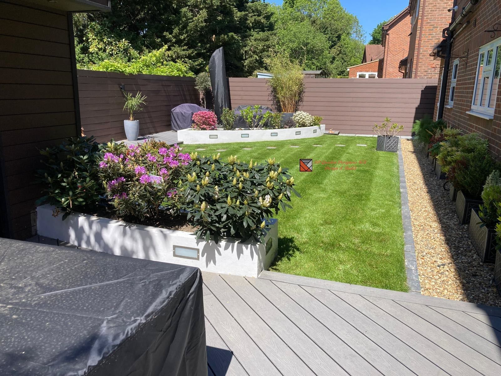A well-manicured garden with a lawn, flowerbeds, a composite deck, and a decorative stone feature, bordered by a wooden fence.