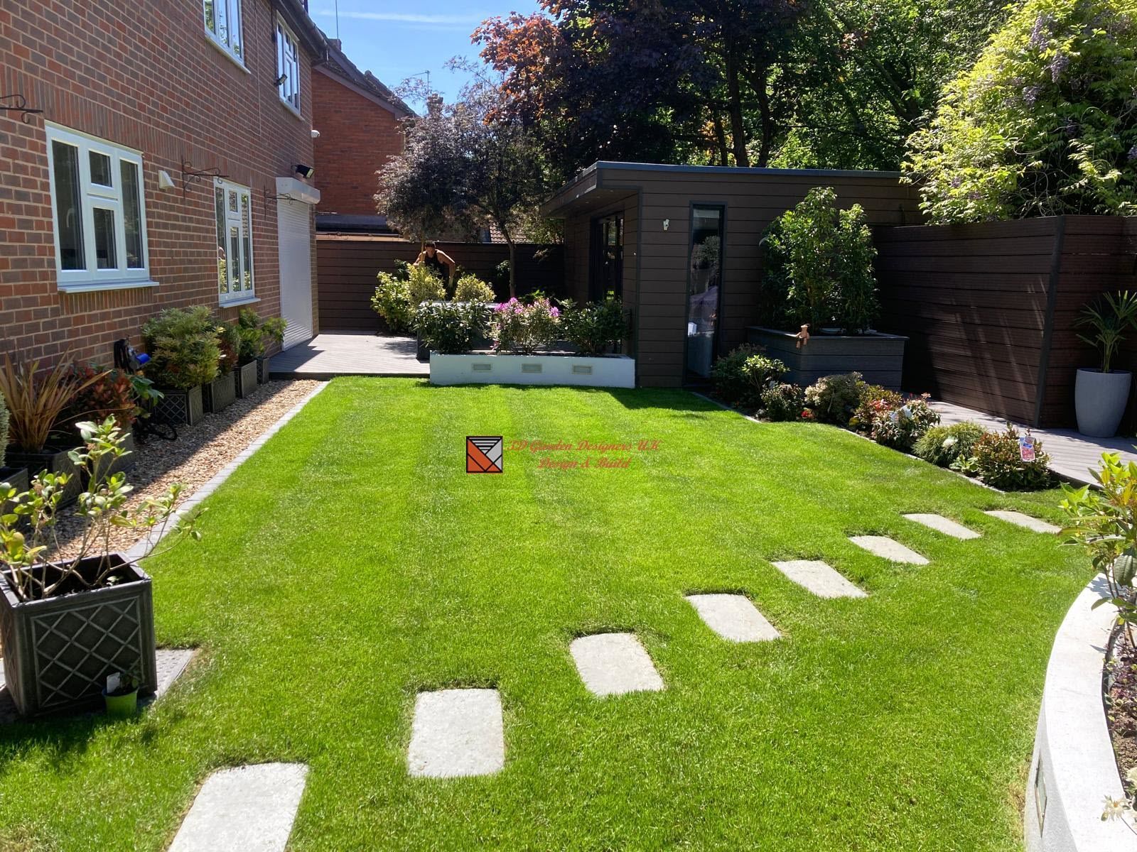 Well-manicured backyard with a green lawn, stepping stones, a modern shed, and flower beds. A brick house is on the left.