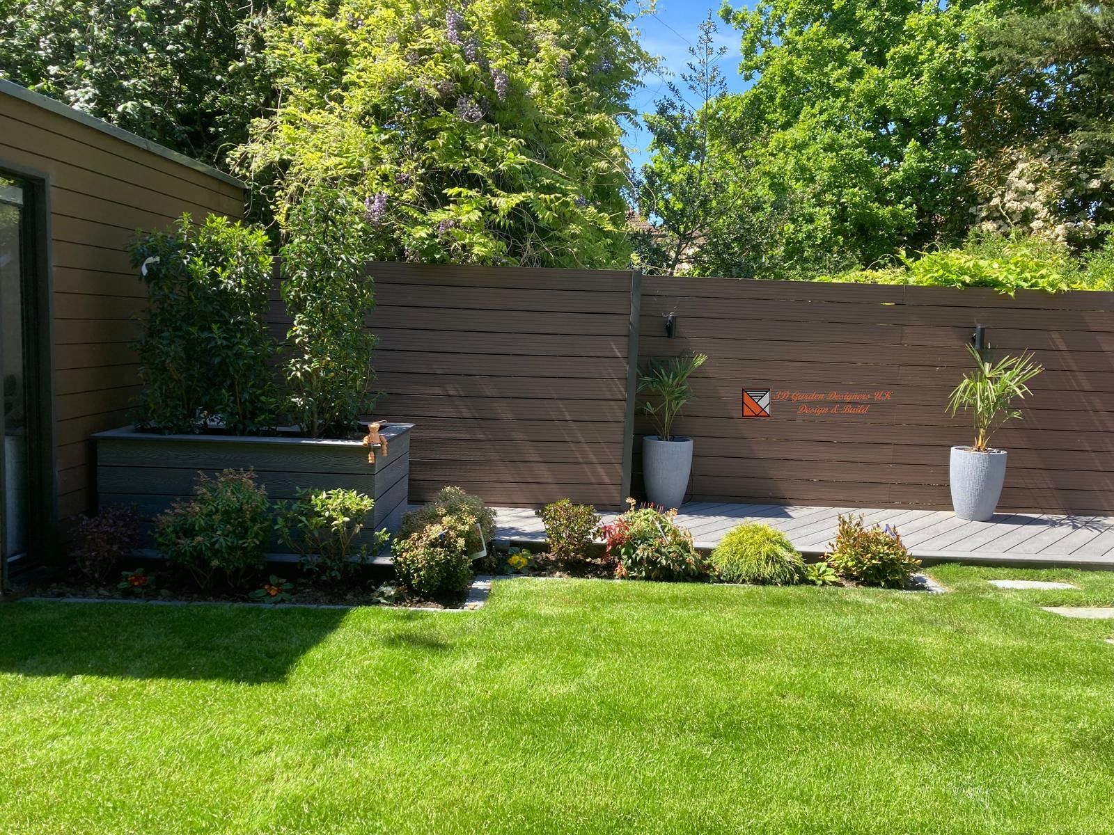 Lush green backyard with a dark brown horizontal slatted fence, planter box, and potted plants. A shed and trees are in the background.