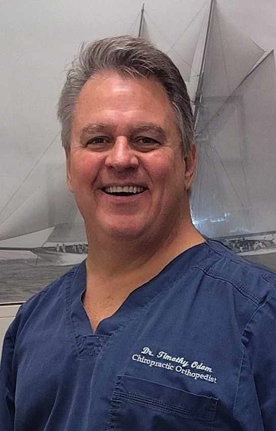 A smiling person in blue scrubs against a blurred background with a sailboat graphic.
