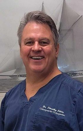 A smiling person in blue scrubs against a blurred background with a sailboat graphic.