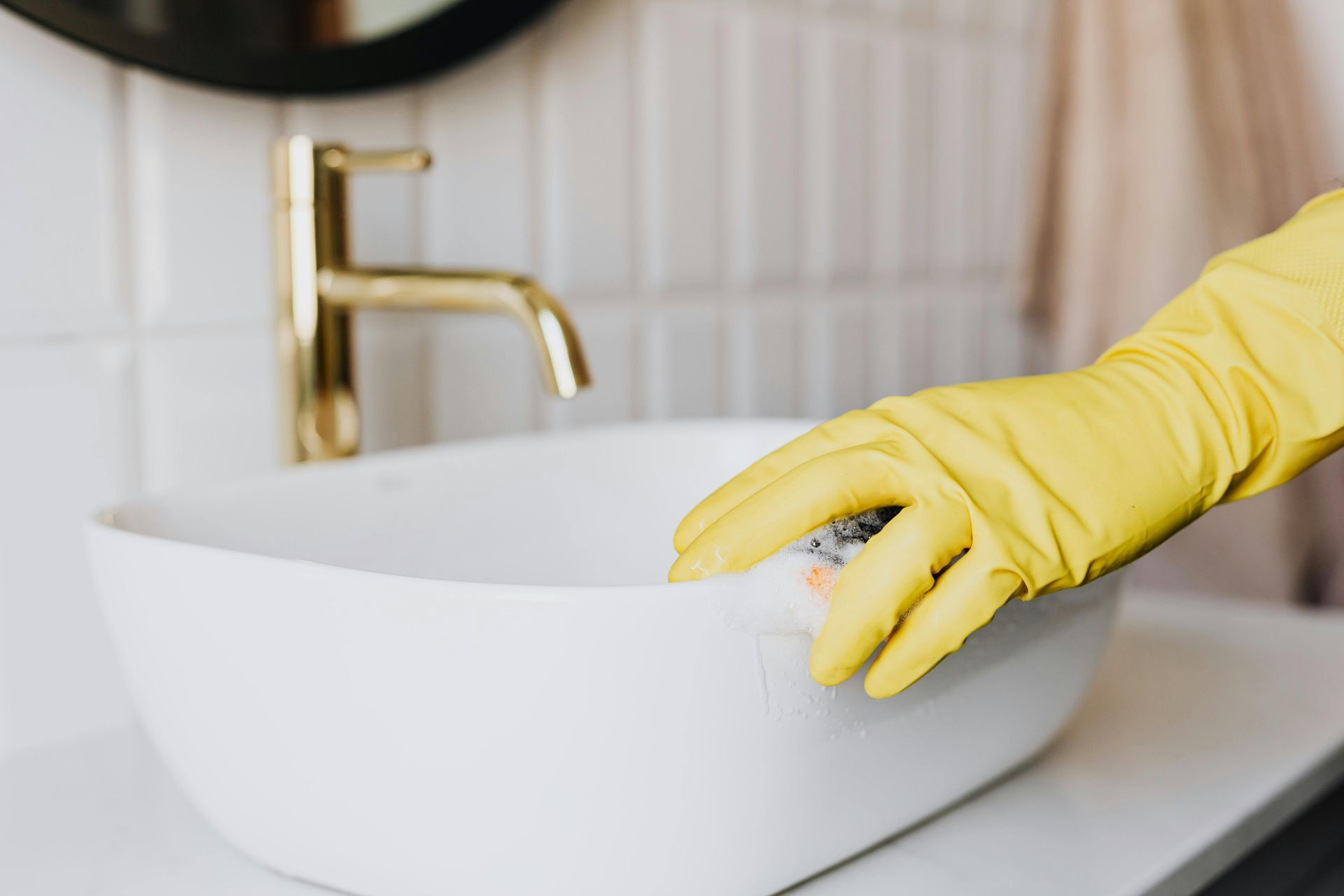 A person wearing yellow gloves is cleaning a bathroom sink with a sponge.