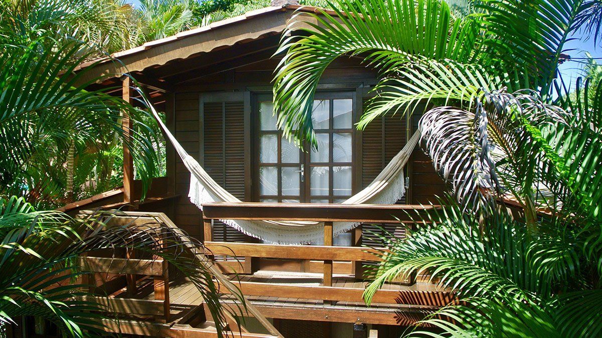 Uma pequena casa de madeira com uma rede na varanda cercada por palmeiras.