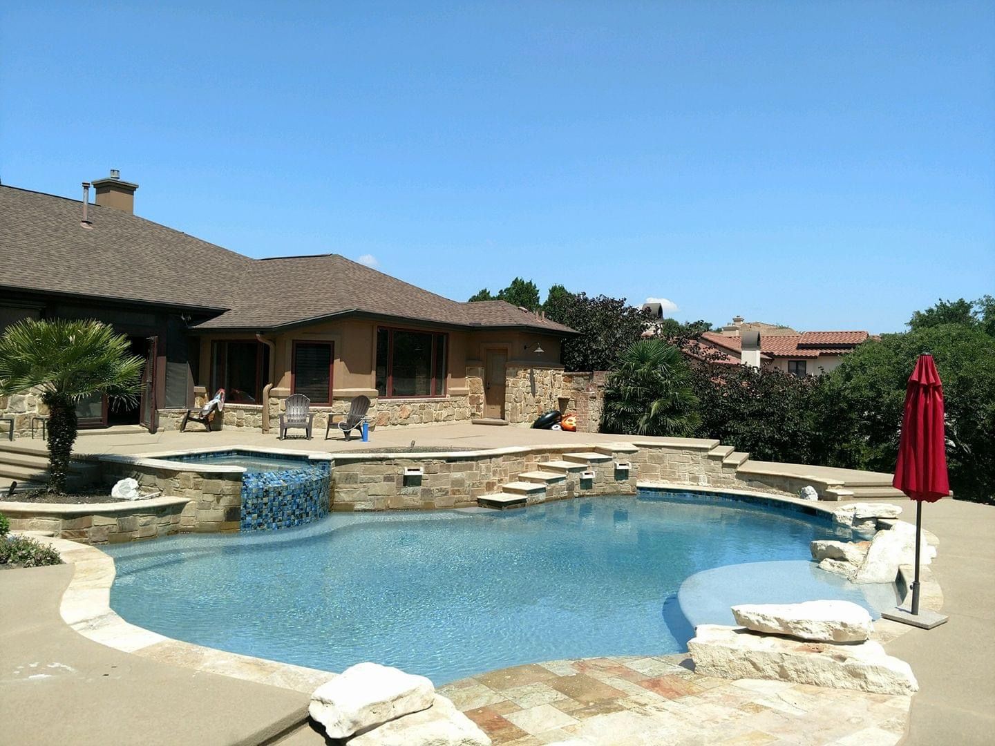 Backyard pool with stone patio, patio furniture, and red umbrella beside a house