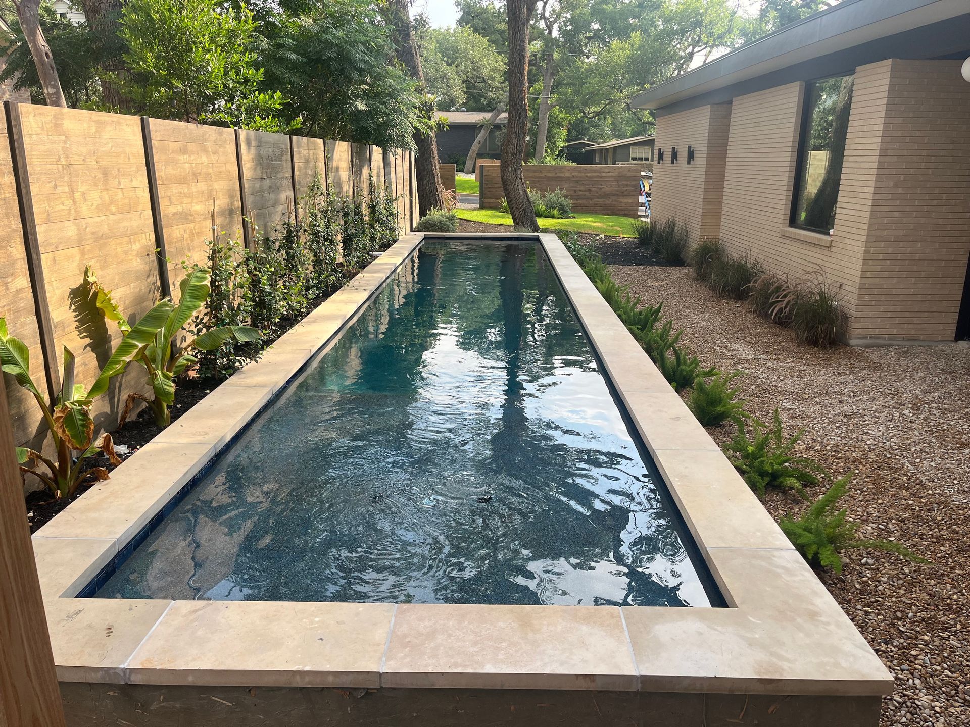 Narrow backyard lap pool with stone coping, bordered by wood fence and greenery beside a house.