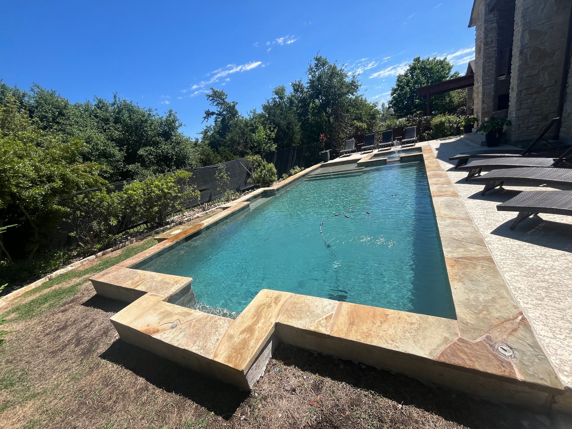 Backyard pool with turquoise water, stone decking, and lounge chairs beside a house under a blue sky