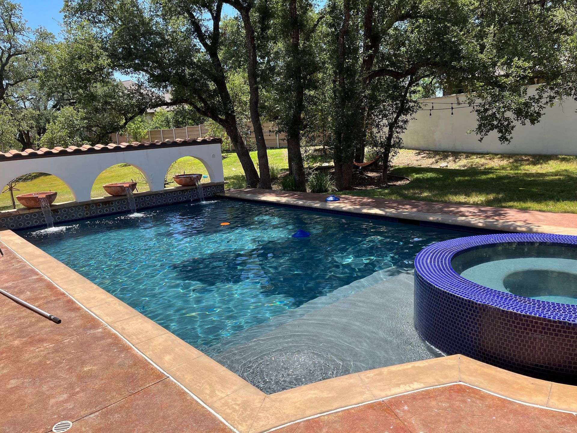 Backyard swimming pool with blue water, stone deck, trees, and a round purple hot tub