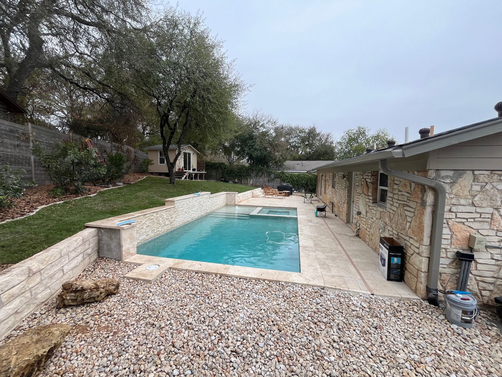 Backyard pool beside a stone house, with gravel, trees, and a cloudy sky
