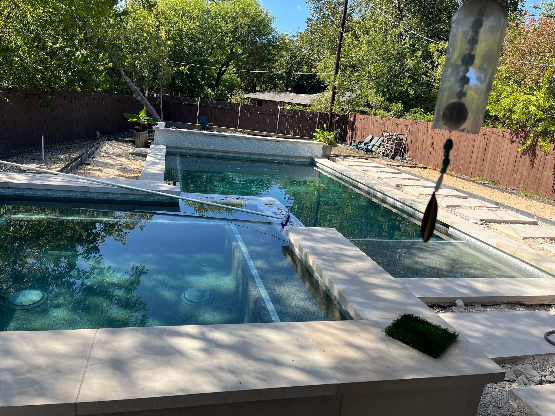 Backyard swimming pool with blue water, patio seating, and trees in bright sunlight