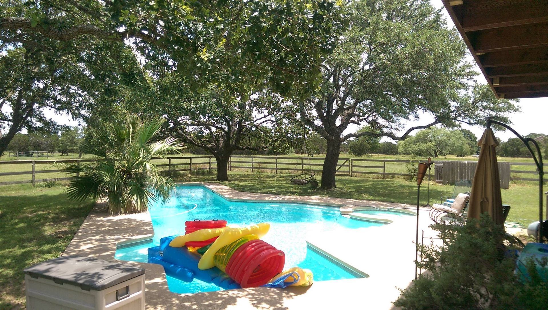 Backyard pool with colorful floaties under trees and a wooden deck overlooking a fenced pasture