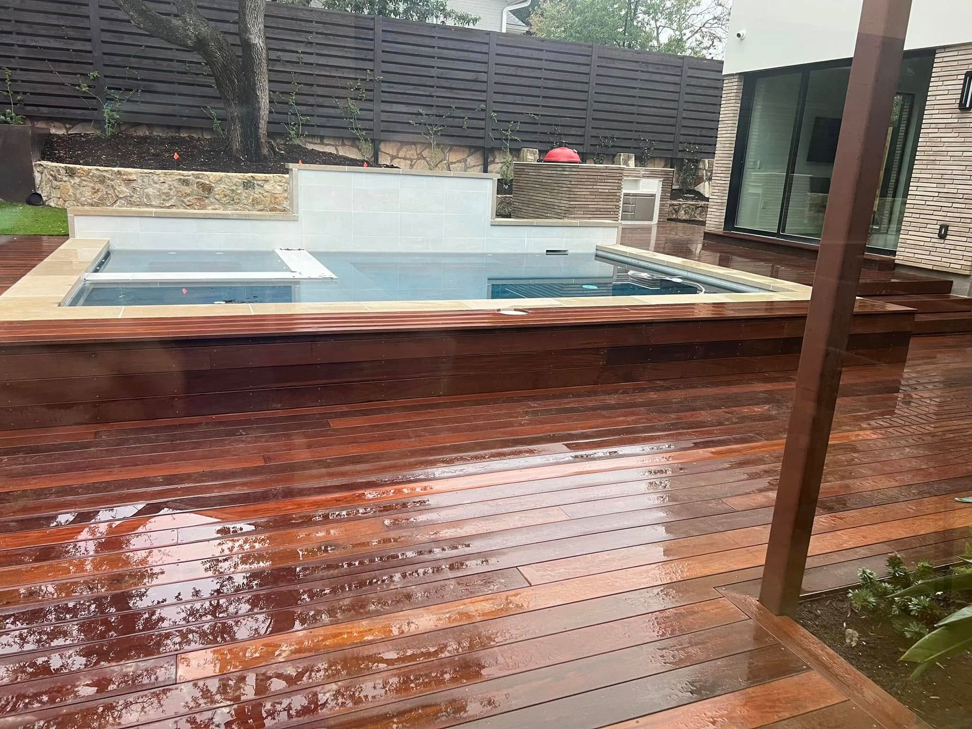 Backyard deck with wet wooden boards beside a small rectangular pool and stone retaining wall.