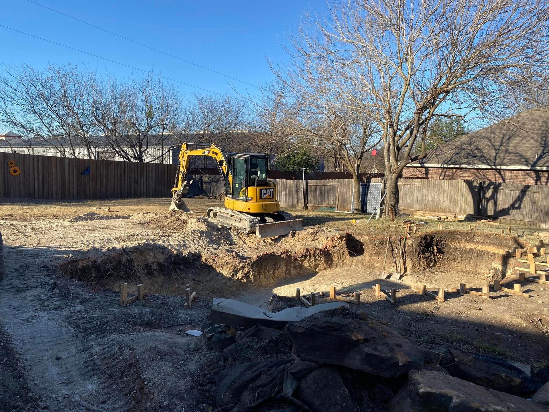 Yellow excavator digging in a backyard with trees and a wooden fence under a clear blue sky
