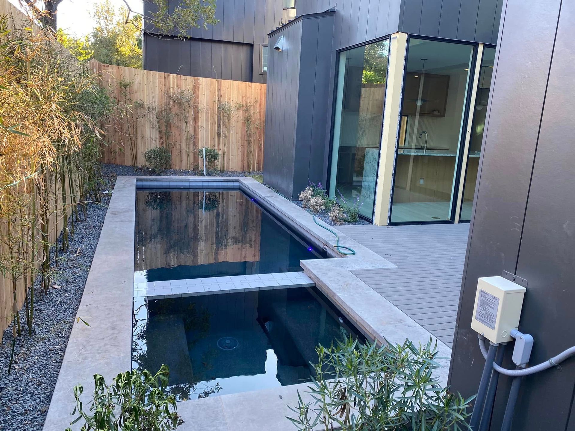 Modern backyard with narrow rectangular pool, dark exterior walls, sliding glass door, and wooden fence.