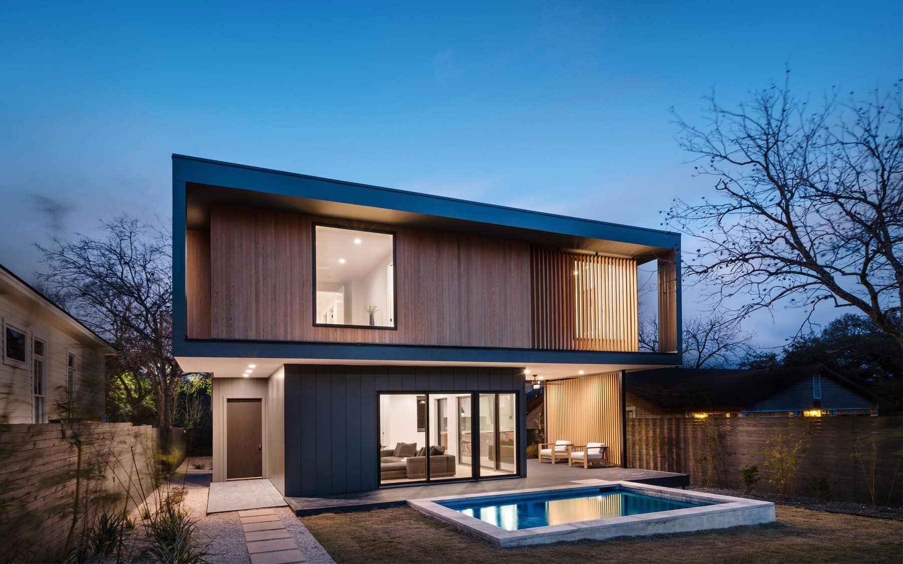 Modern two-story house with large windows and a small lit pool at dusk