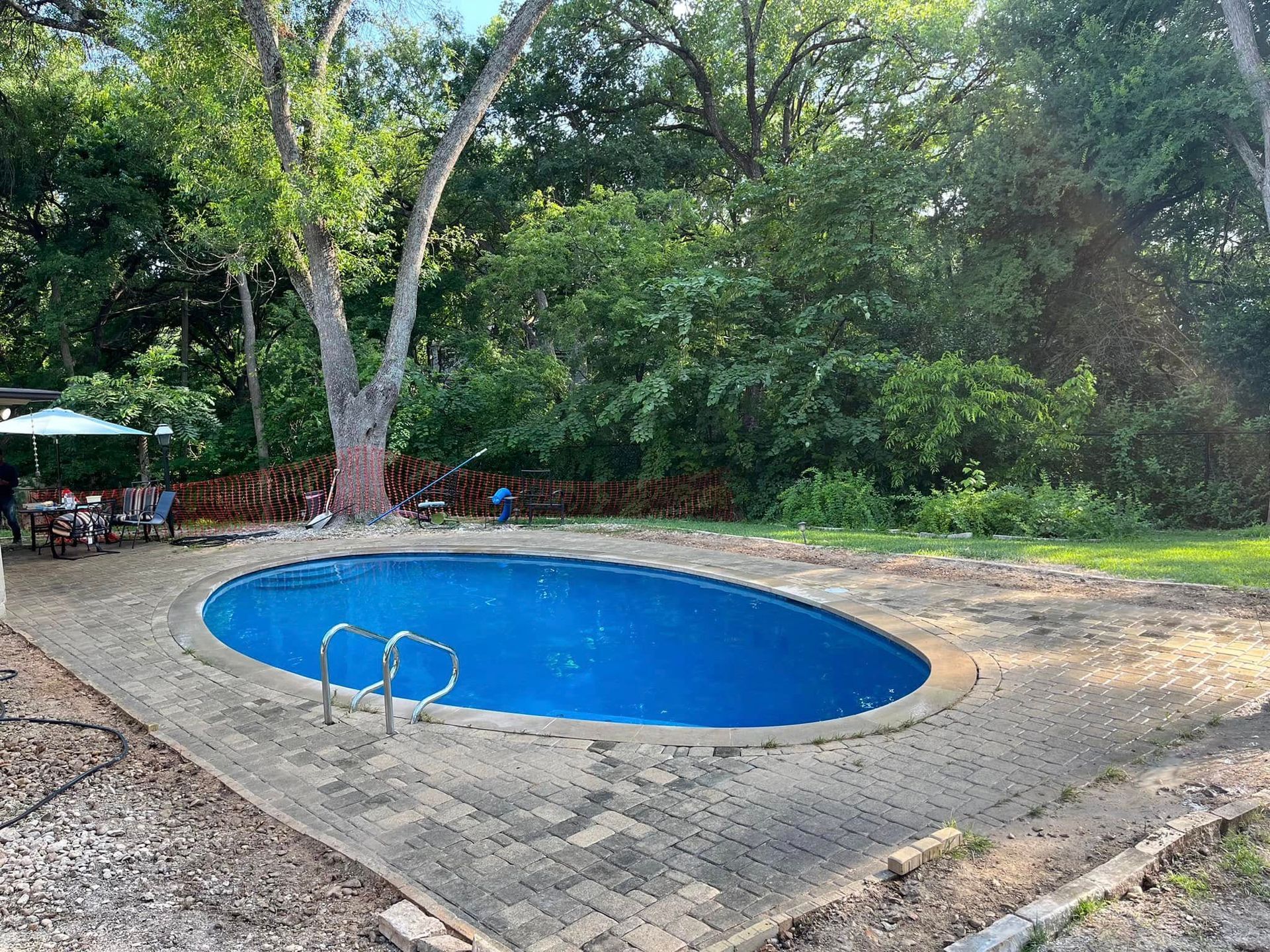 Small blue oval swimming pool surrounded by stone paving and trees in a wooded yard