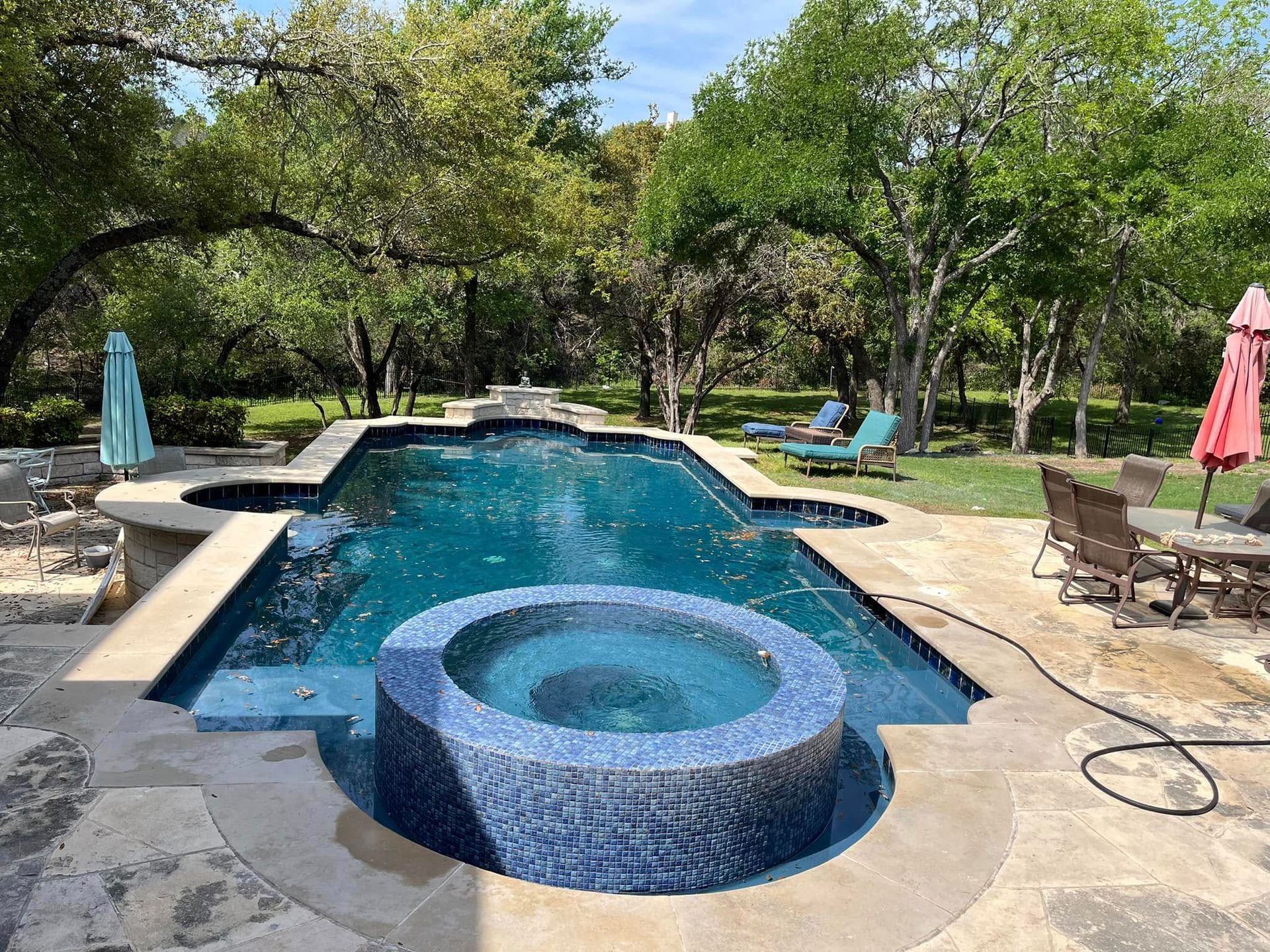 Backyard pool with a round hot tub, stone deck, lounge chairs, and umbrellas under trees