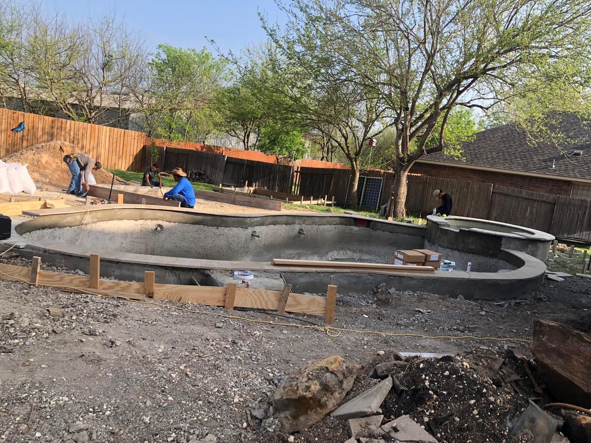 Workers building a circular concrete structure in a dusty yard with trees and a house nearby