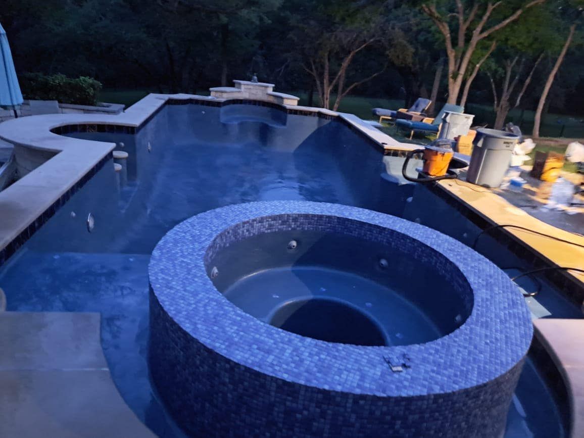 Backyard pool at dusk with a round spa in the foreground and pool equipment on the deck.