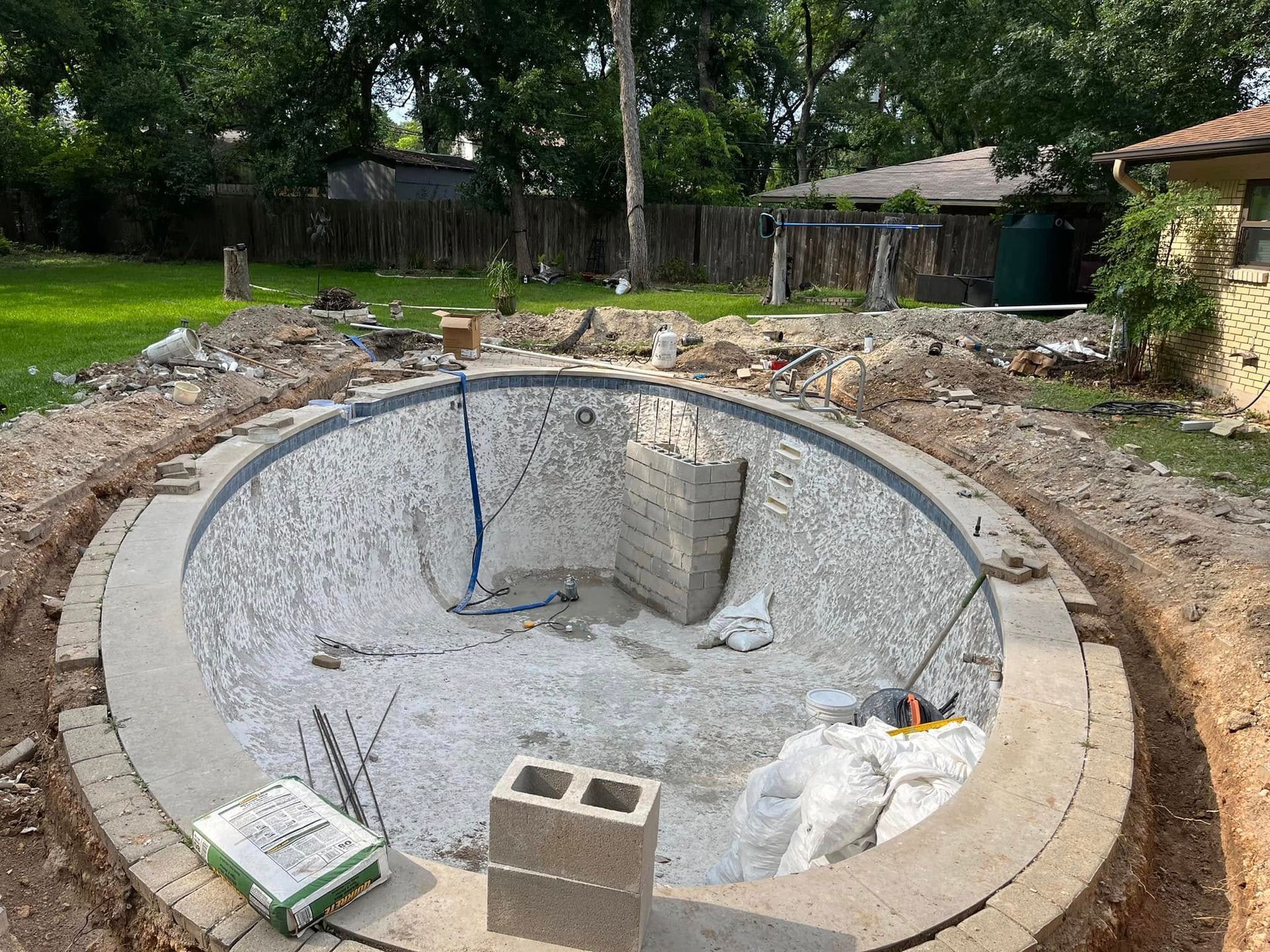Empty backyard pool under construction with exposed liner, concrete coping, and scattered materials