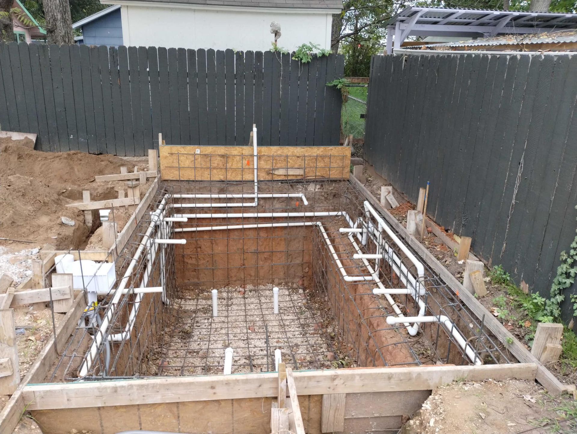 Backyard pool excavation with rebar and plumbing forms inside a wooden frame, bordered by dark fences