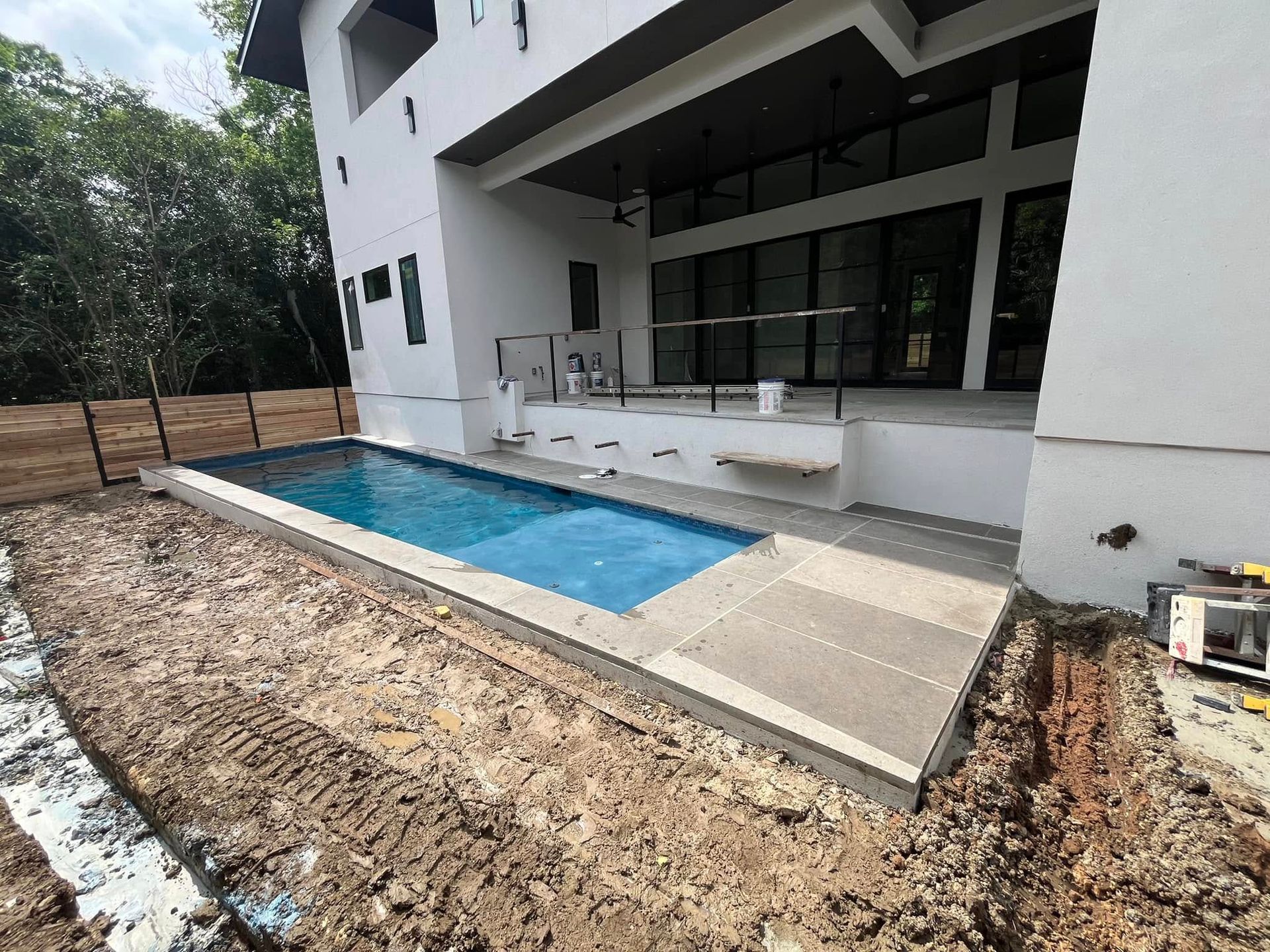 Modern house patio with a rectangular blue pool and concrete deck, surrounded by dirt landscaping.